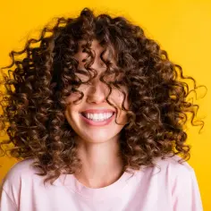 Uśmiechnięta kobieta z burzą brązowych, kręconych włosów (coily hair) na tle żółtej ściany.